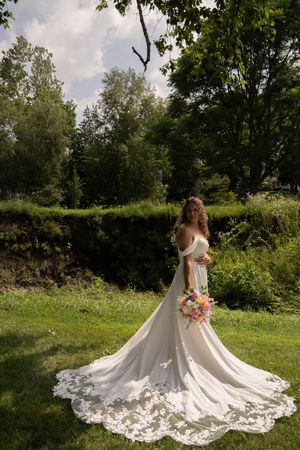 A stunning white wedding dress with lace train spreads across a lush green meadow on a sunny summer day.