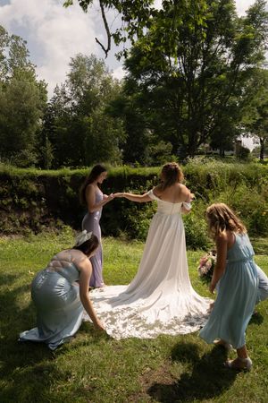 Bridesmaids in pastel dresses help arrange the bride's flowing white train on grassy ground.