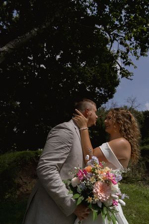 Romantic outdoor wedding portrait with pink floral bouquet against lush greenery.
