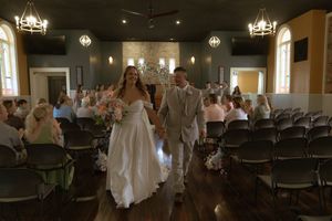 A newlywed couple walks down the aisle in a chapel with wooden chairs and hanging lights.