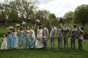 Wedding party in light blue dresses and gray suits poses together on a grassy lawn under cloudy skies.