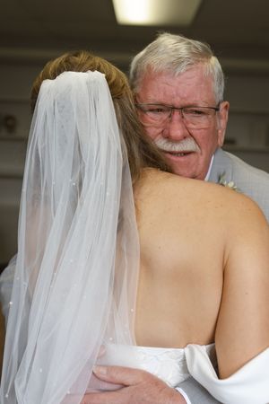 A bride in a white wedding dress shares a touching moment with an older gentleman wearing glasses.