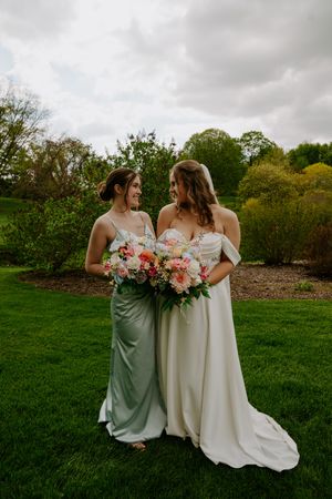 Bridesmaids in sage green dresses pose with bride in white gown holding pink and coral bouquets.