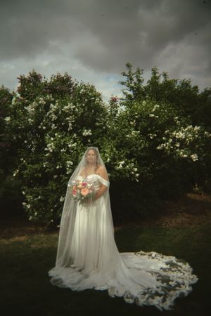 Dramatic bridal portrait in white gown and flowing veil against flowering garden backdrop.