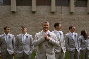 Wedding party in coordinated light gray suits poses against stone wall exterior.