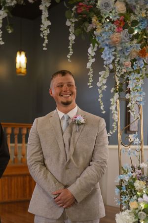Wedding ceremony setup with floral arch and groom in beige suit standing underneath.