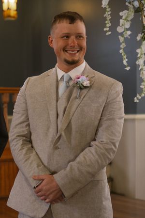 A groom in a light beige suit with a pink boutonniere smiles during a wedding ceremony.