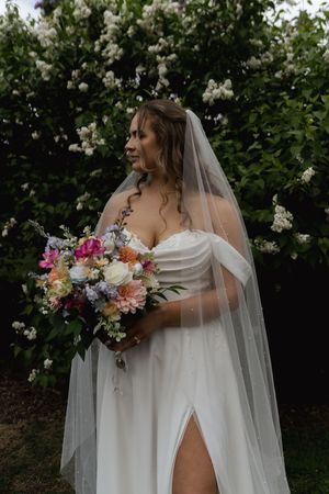 Bride in off-shoulder wedding dress holds colorful bouquet against blooming white flowers.