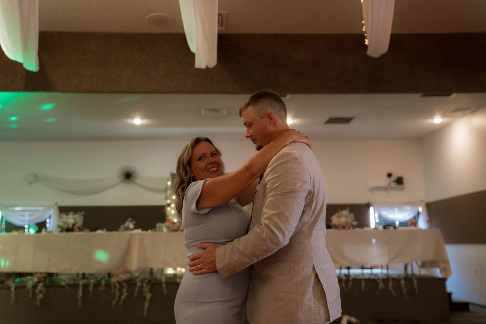 A tender slow dance moment in a reception hall with decorative lights and white draping.