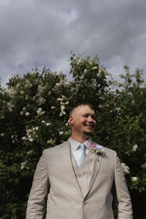 Elegant groomsman in light gray suit and blue tie poses against flowering bush backdrop.