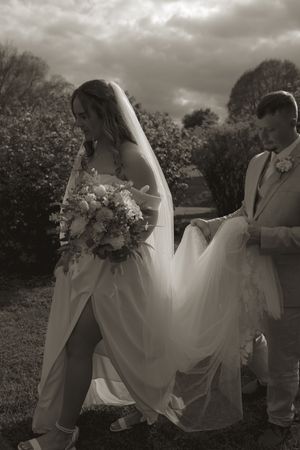 A bride in a flowing white gown holds a bouquet while her dress is adjusted in a garden setting.