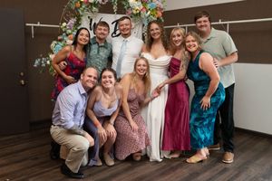 Group photo of wedding guests seated on chairs in a reception venue with elegant draping and chandeliers.