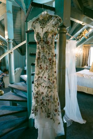 Delicate floral wedding dress hanging on turquoise staircase with romantic lighting.