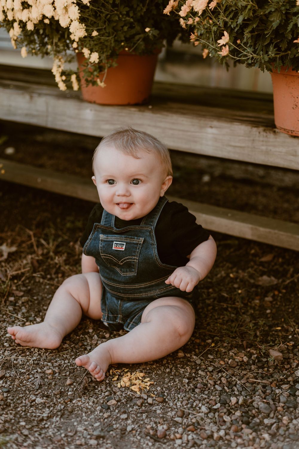 An adorable baby in denim overalls sits barefoot on gravel beside potted flowers outdoors.
