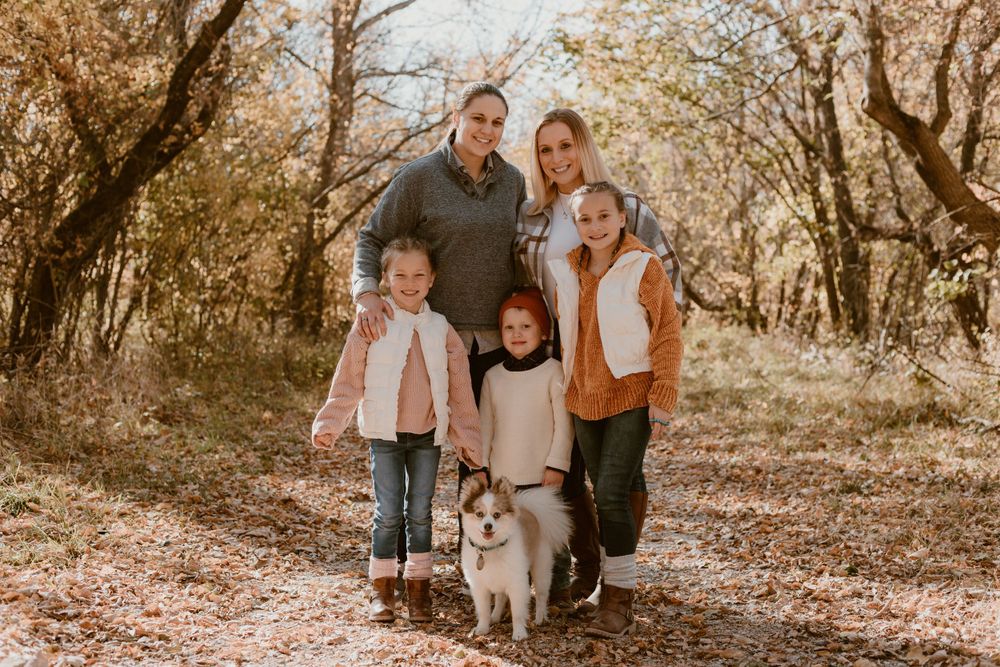 A happy family of five with a corgi dog poses on a leaf-covered path in fall woods.