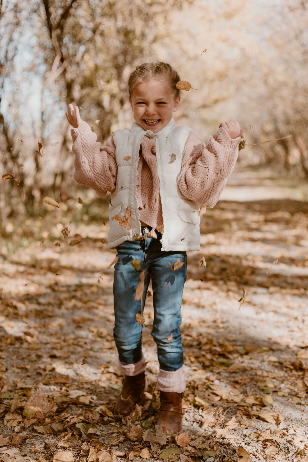 A joyful young girl tosses autumn leaves in a sunlit forest, wearing a pink sweater and jeans.