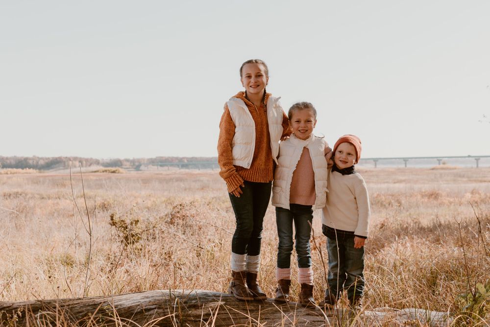 Three sisters pose together on a log in a golden autumn field under a clear sky.
