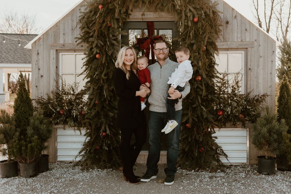 Smiling couple holding two babies standing under Christmas garland arch outside rustic farmhouse.