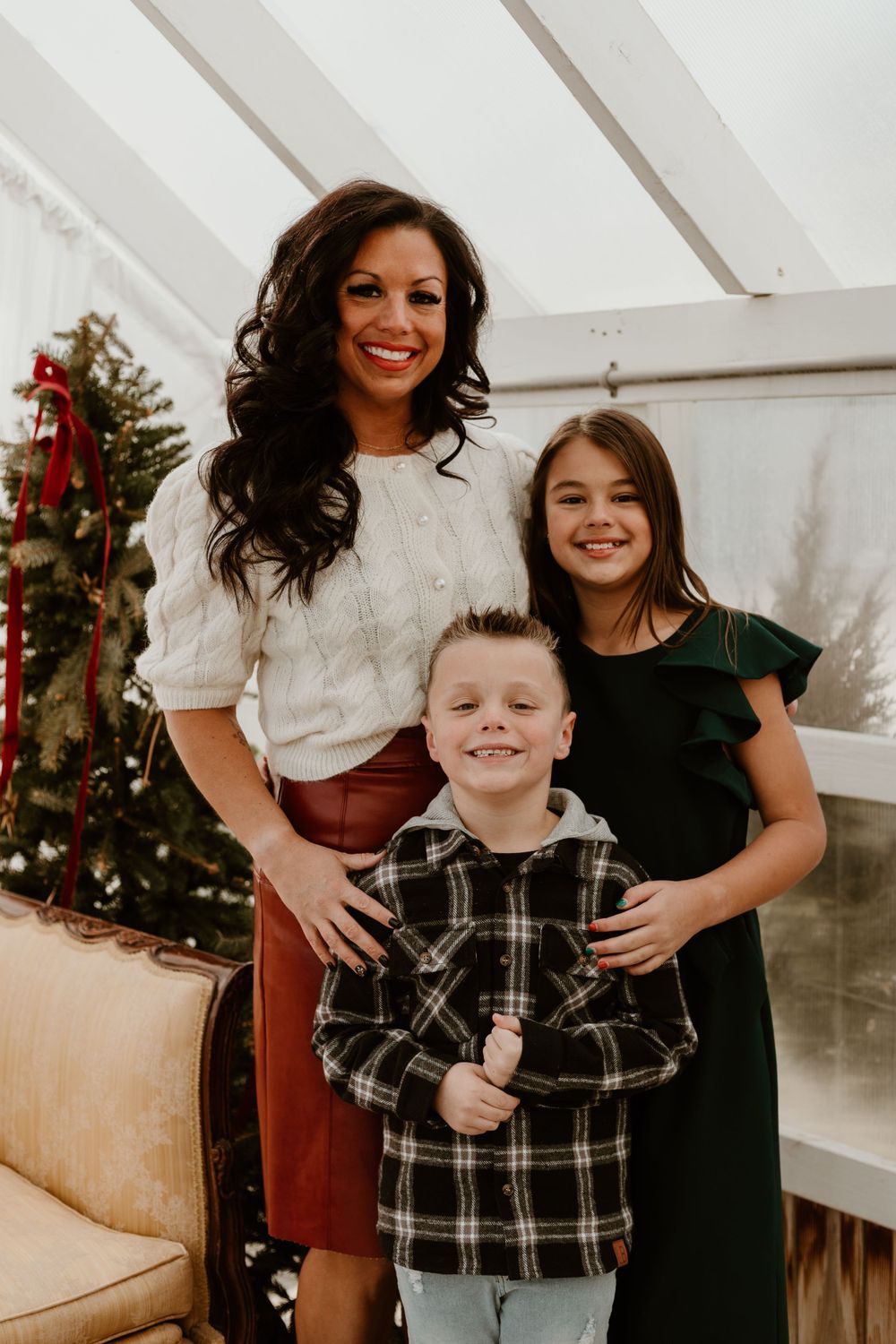 Smiling woman with two children posing in front of Christmas tree in festive holiday setting.