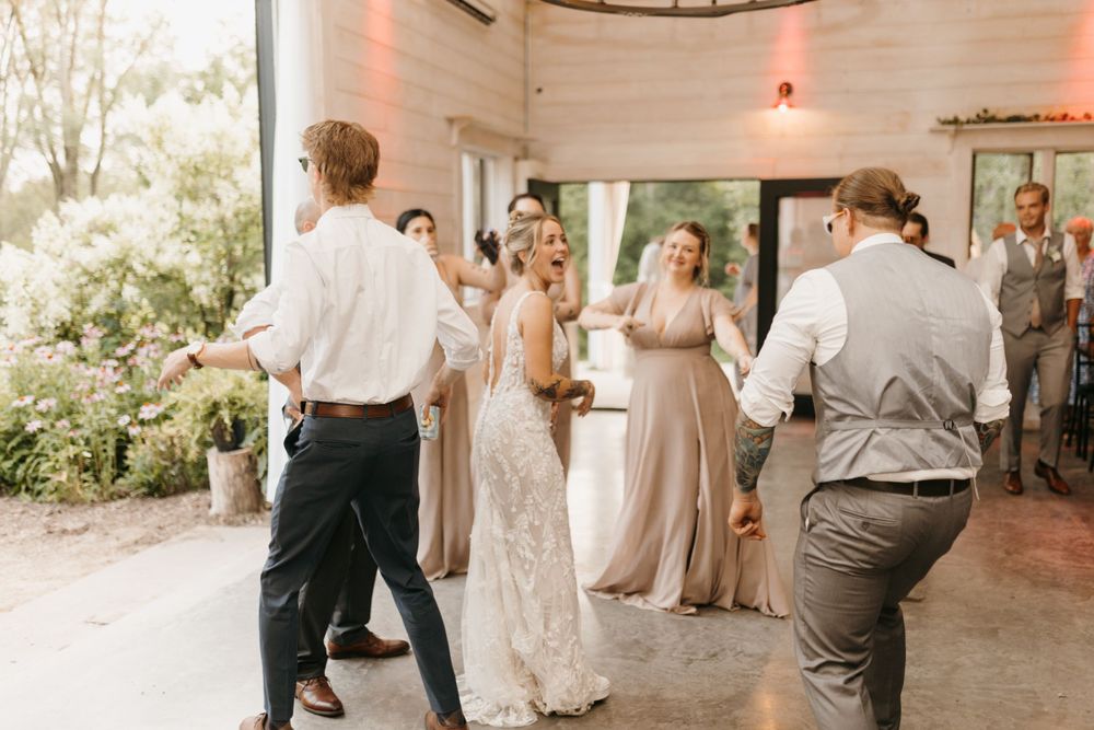 Bride in white gown dances joyfully with bridesmaids in taupe dresses at reception.