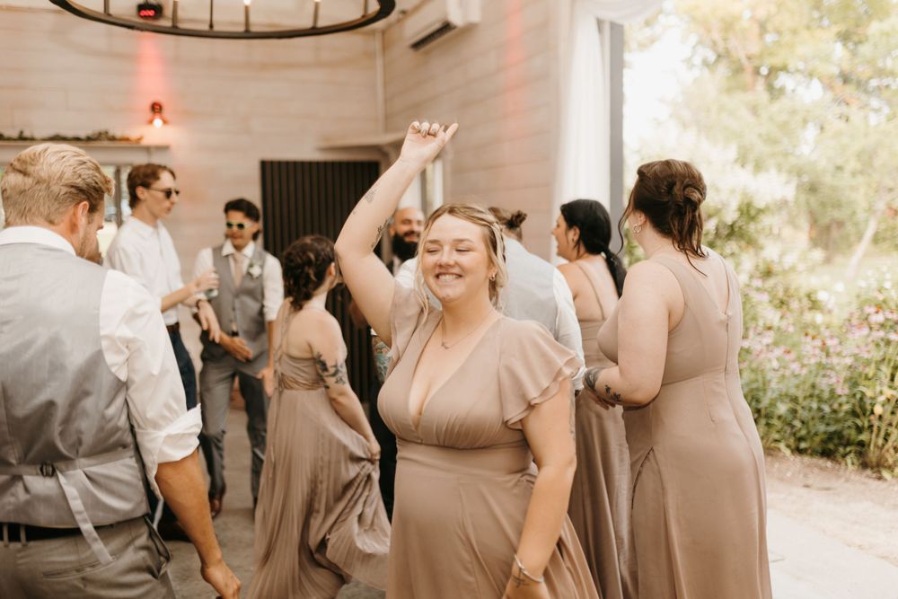 Bridesmaid in taupe dress dances with arm raised joyfully at wedding reception.