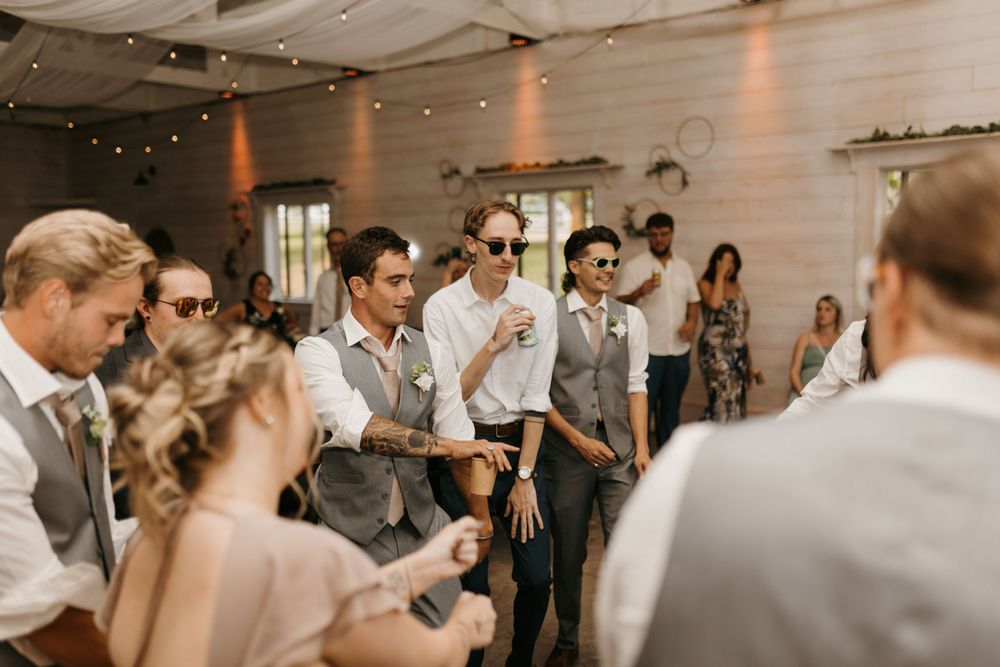 Groomsmen in vests and sunglasses dance together at a rustic barn wedding reception.