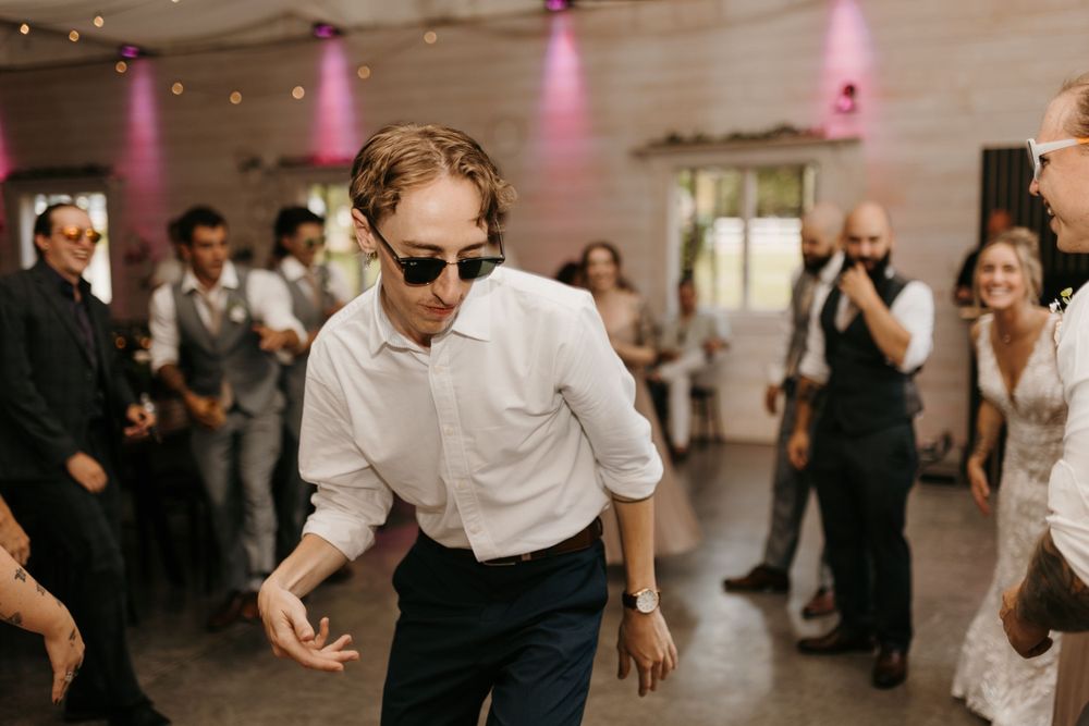 A man in sunglasses and white shirt dances energetically at a wedding reception.