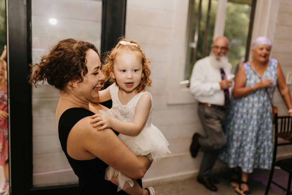 A woman holds a curly-haired toddler in a white dress at a wedding reception.