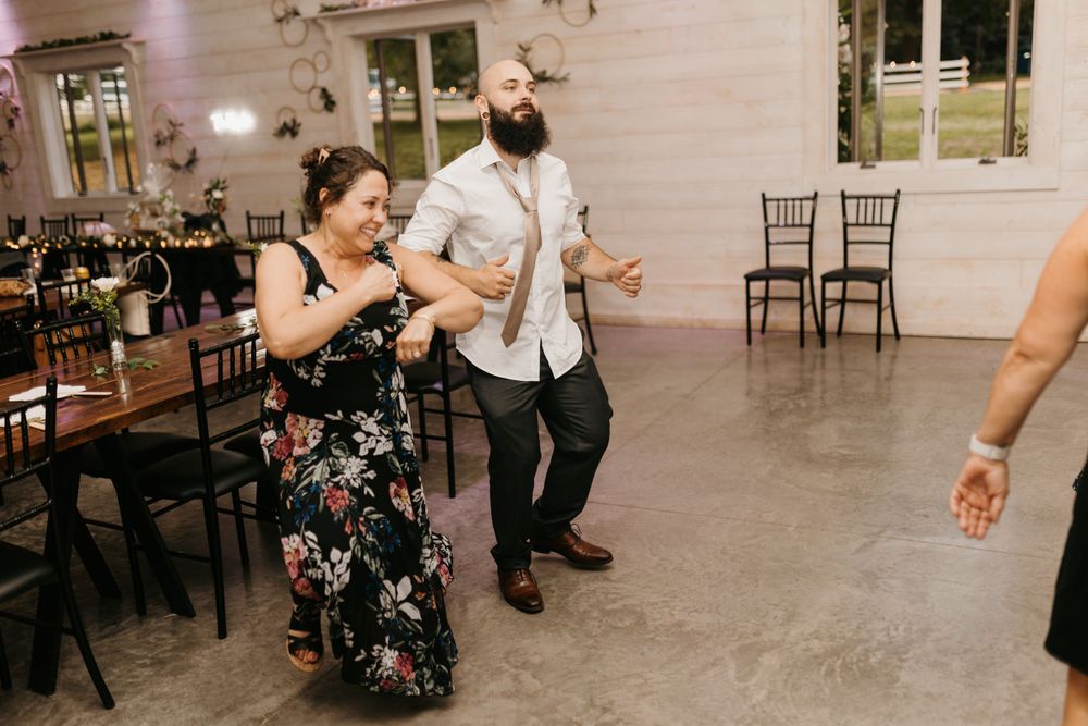 A bearded man and woman in a floral dress dance joyfully at a rustic wedding reception venue.