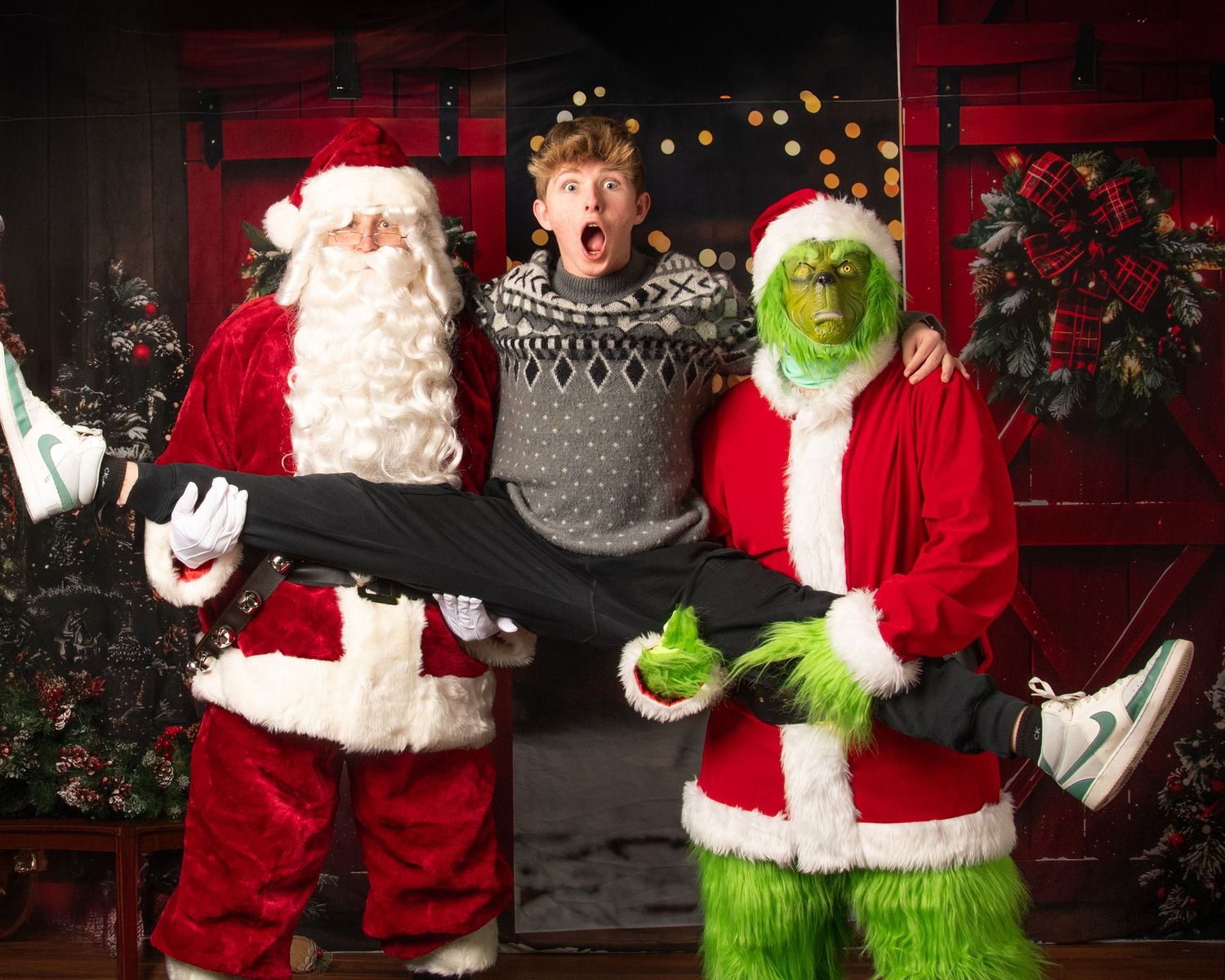 Holiday photo featuring Santa and the Grinch posing with someone in a patterned sweater against decorated backdrop.