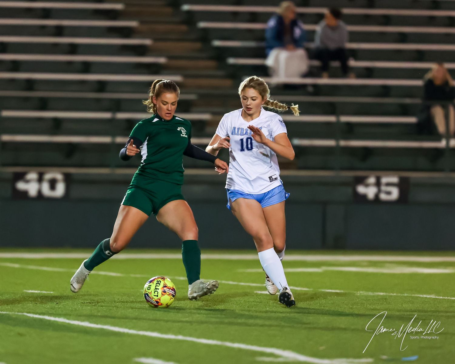 Two women's soccer players compete for the ball during a night game, one in green and one in white uniforms.