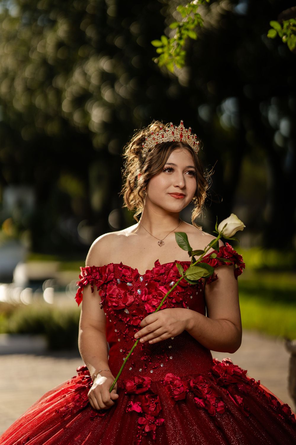 A stunning red ball gown with floral details and a white rose is worn in a dreamy outdoor setting.