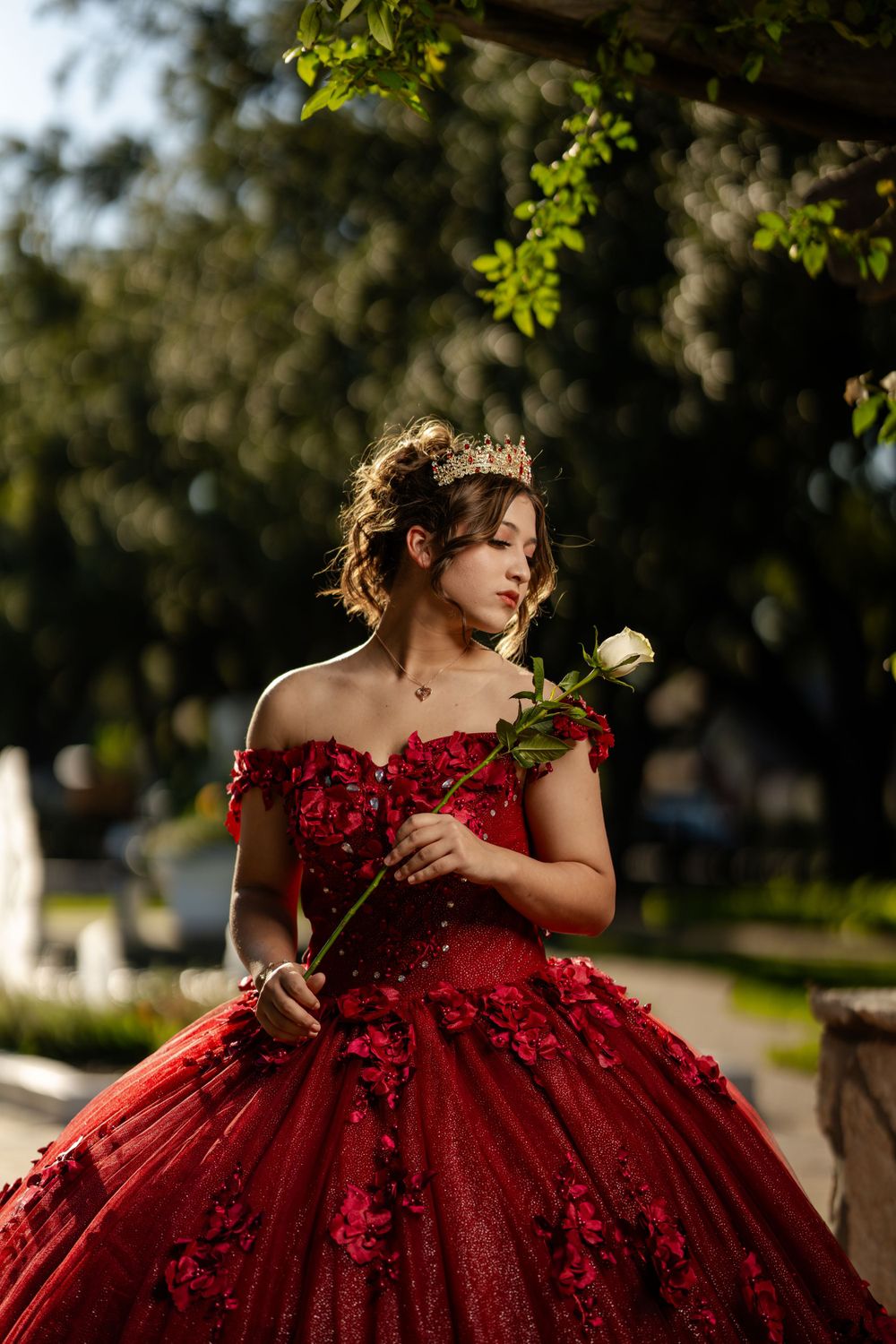 A person in a stunning red ball gown with floral details holds a white rose while standing in a sunlit garden setting.