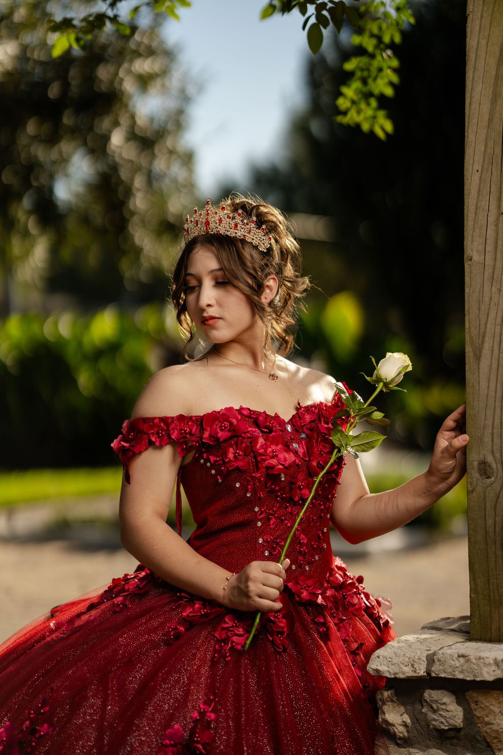A person in an elegant red off-shoulder ball gown holds a white rose while posing outdoors in natural lighting.