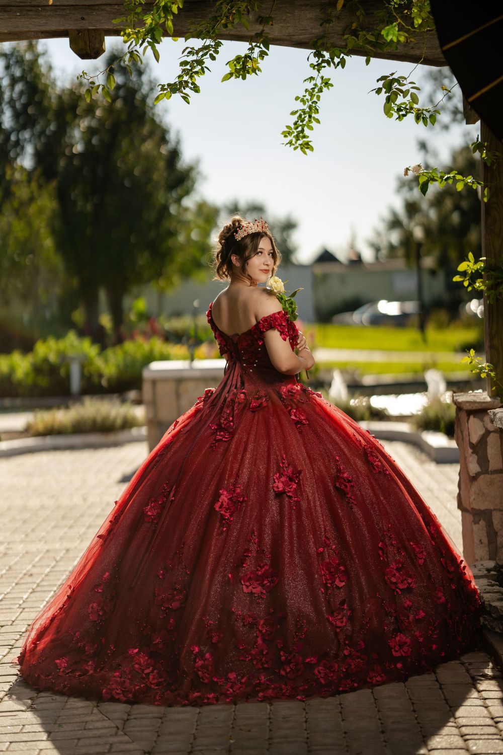 Elegant burgundy ball gown with floral appliques photographed in a garden setting with natural sunlight.