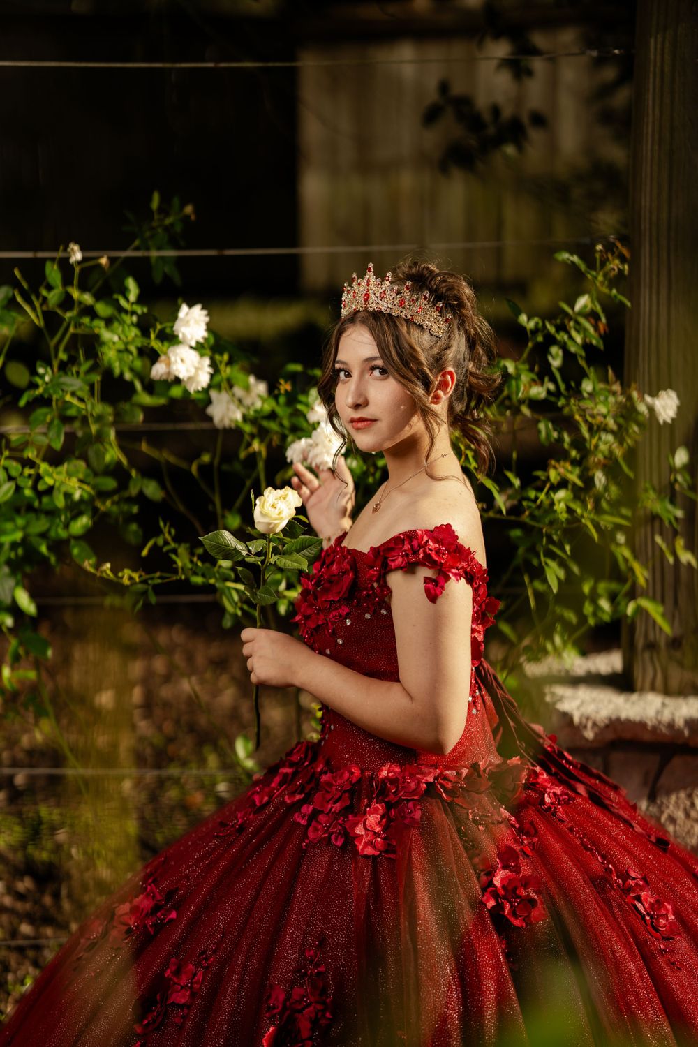 A person in an ornate red ball gown and crown poses elegantly among white roses in a garden setting.