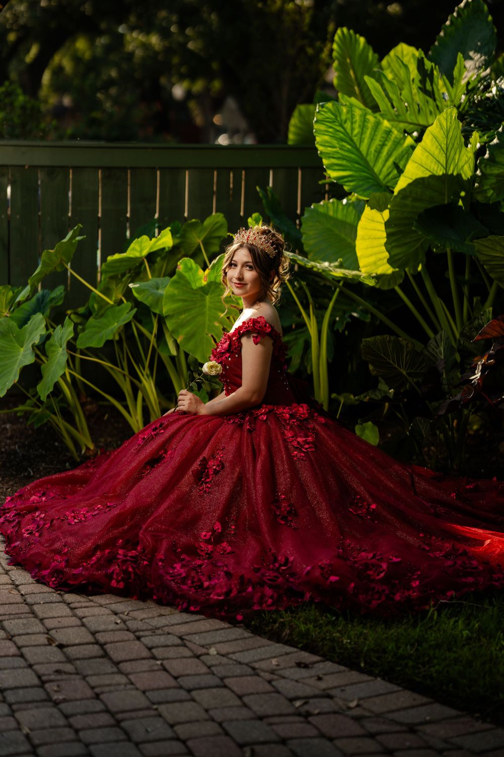 A stunning red ball gown with intricate details photographed in a garden setting with large tropical leaves.