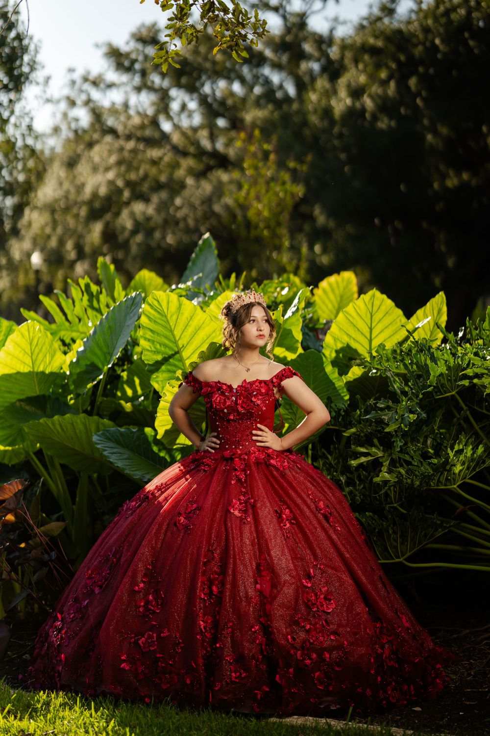 A stunning deep red ball gown with floral details photographed in a lush garden setting with large tropical leaves.