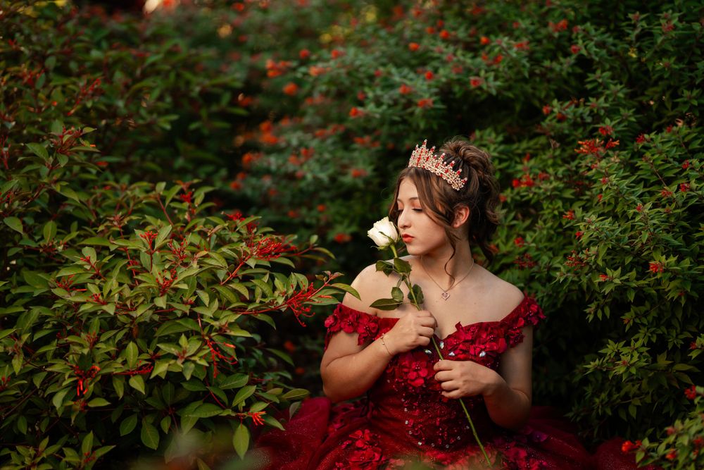 A figure in a red sequined gown and crown sits among lush greenery holding a white rose in a dreamy garden setting.