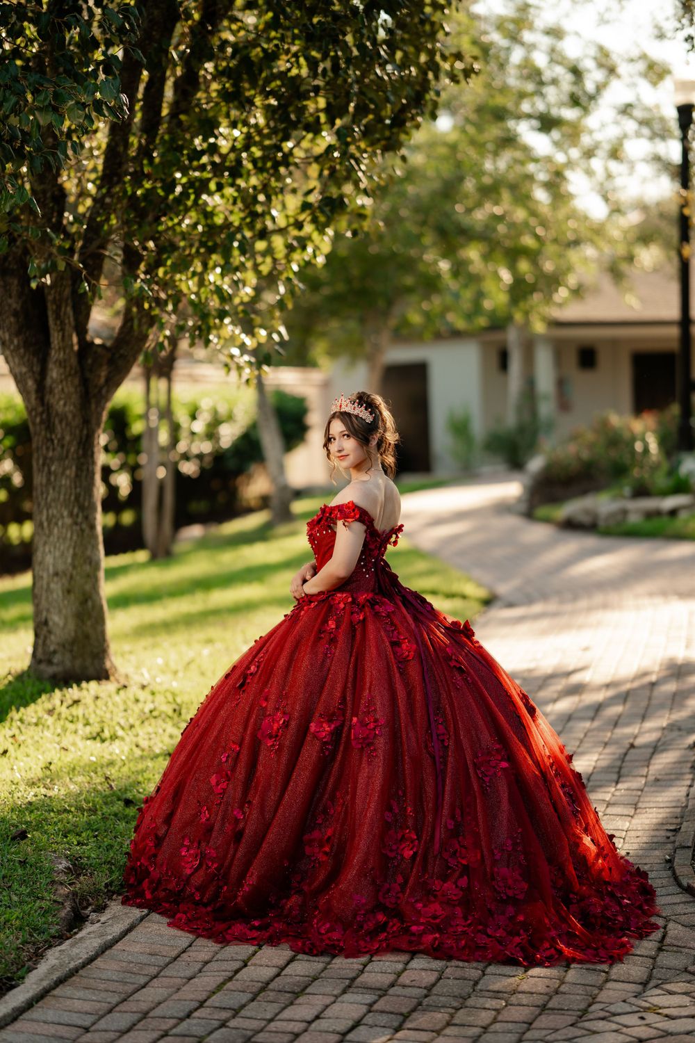 A stunning red ball gown with off-shoulder design photographed on a brick path under trees in natural sunlight.