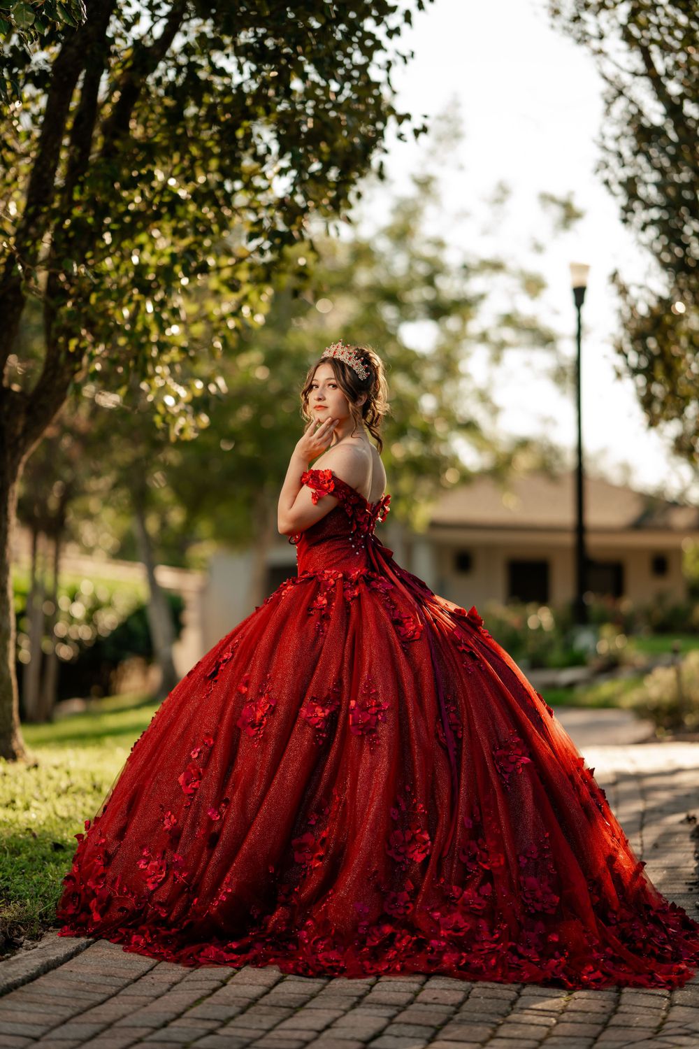 A stunning red ball gown with floral embellishments photographed in a garden setting with natural sunlight filtering through trees.