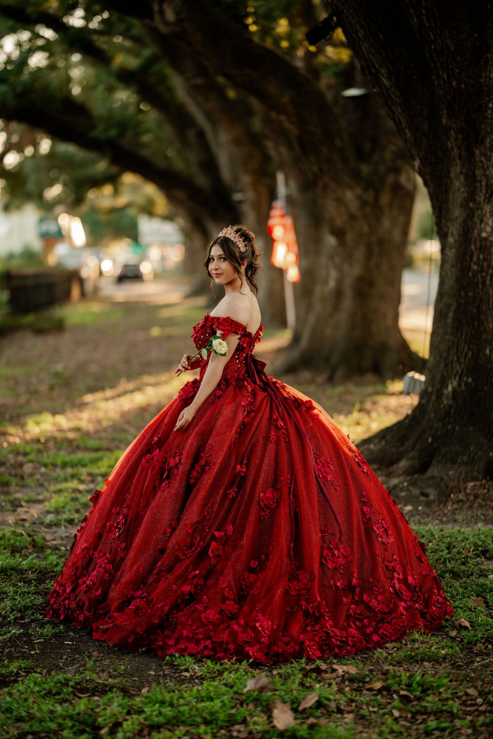 A stunning red ball gown with floral details photographed in a dreamy outdoor setting with large trees in the background.