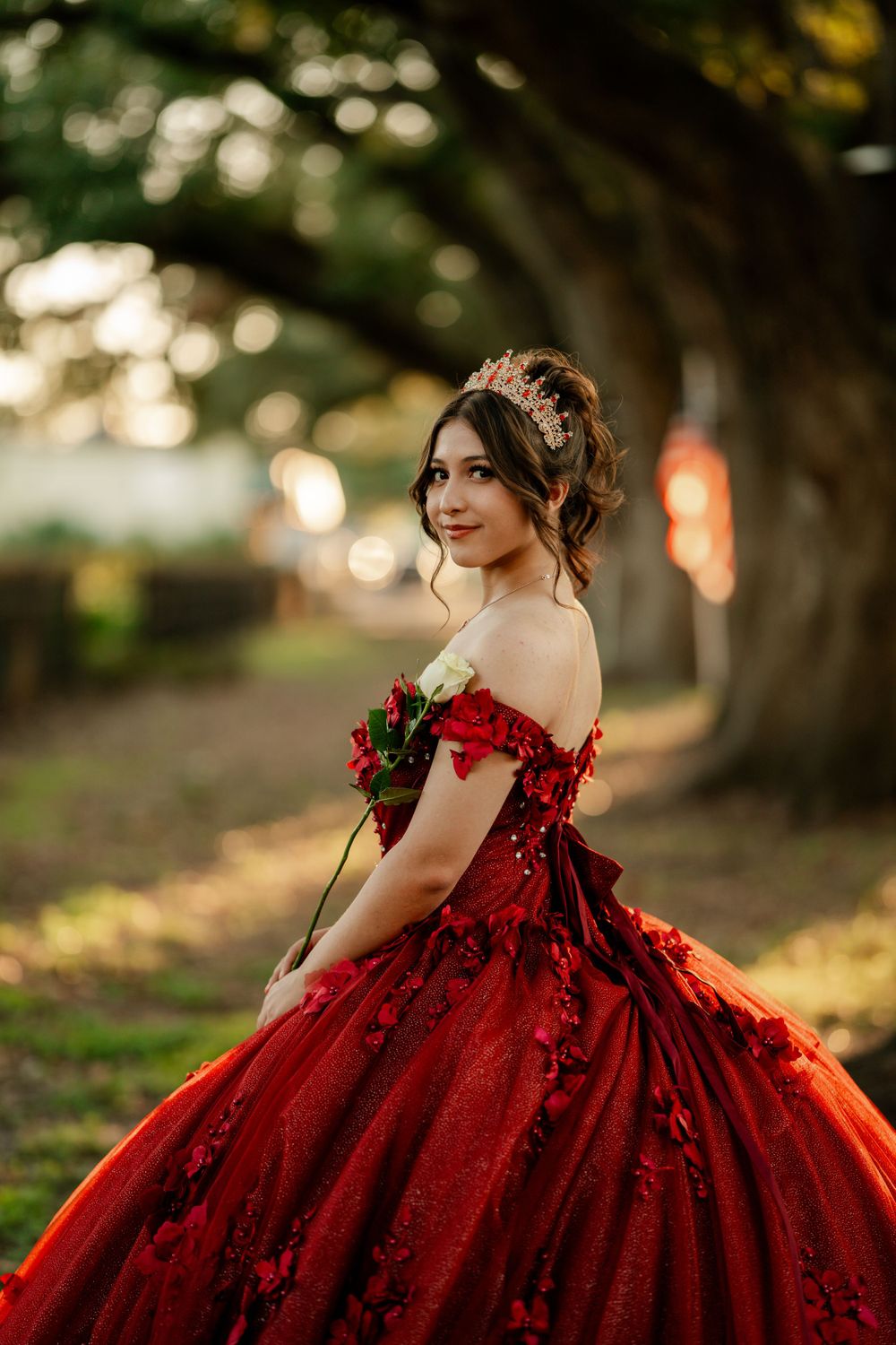 A stunning red ball gown with off-shoulder details photographed in a dreamy outdoor setting with bokeh lighting.