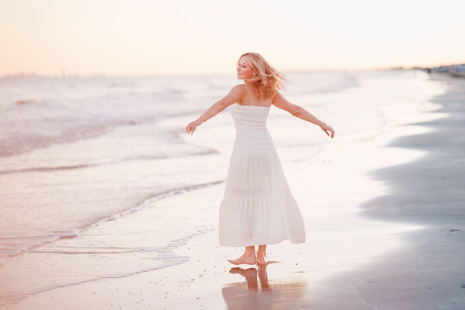 Couple holding hands walking barefoot on a reflective beach at sunset, woman in jeans looking back over shoulder.