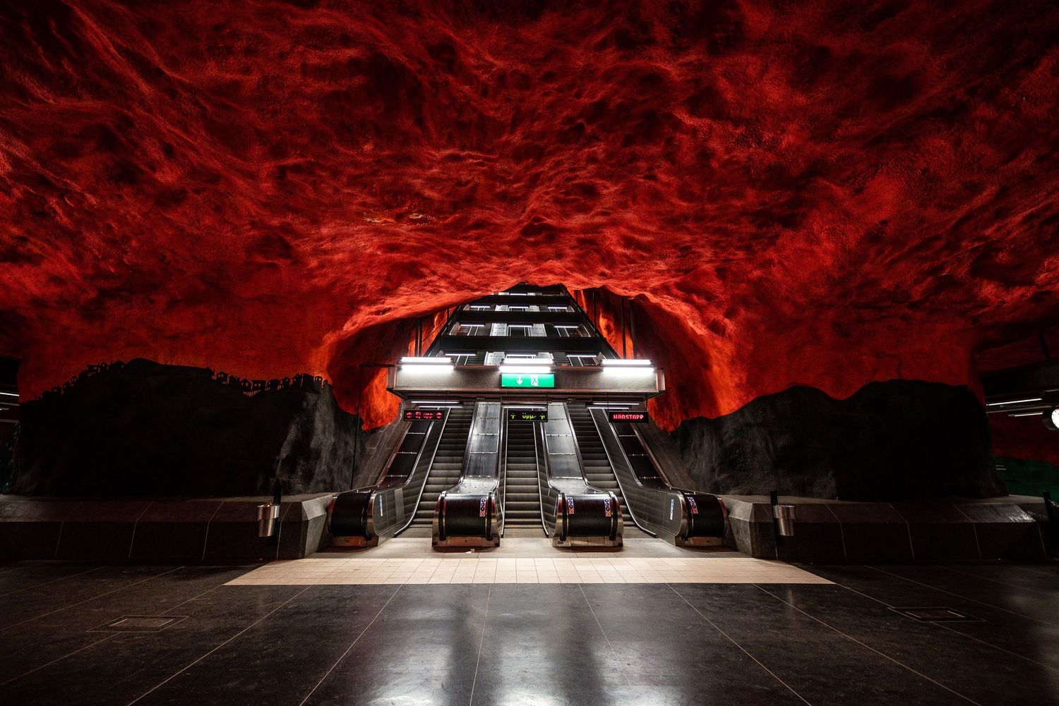 Dramatic red-lit subway station entrance with escalators descending into a cave-like atmosphere. Stockholm metro station