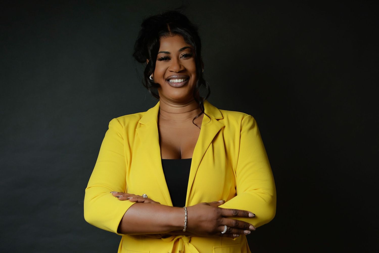 Confident woman in yellow blazer smiling with arms crossed against dark background.
