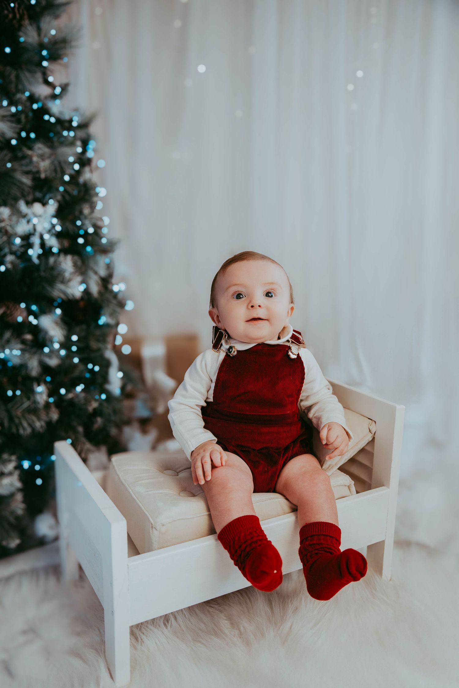 Baby wearing red velvet overalls sits on small white bench beside Christmas tree.