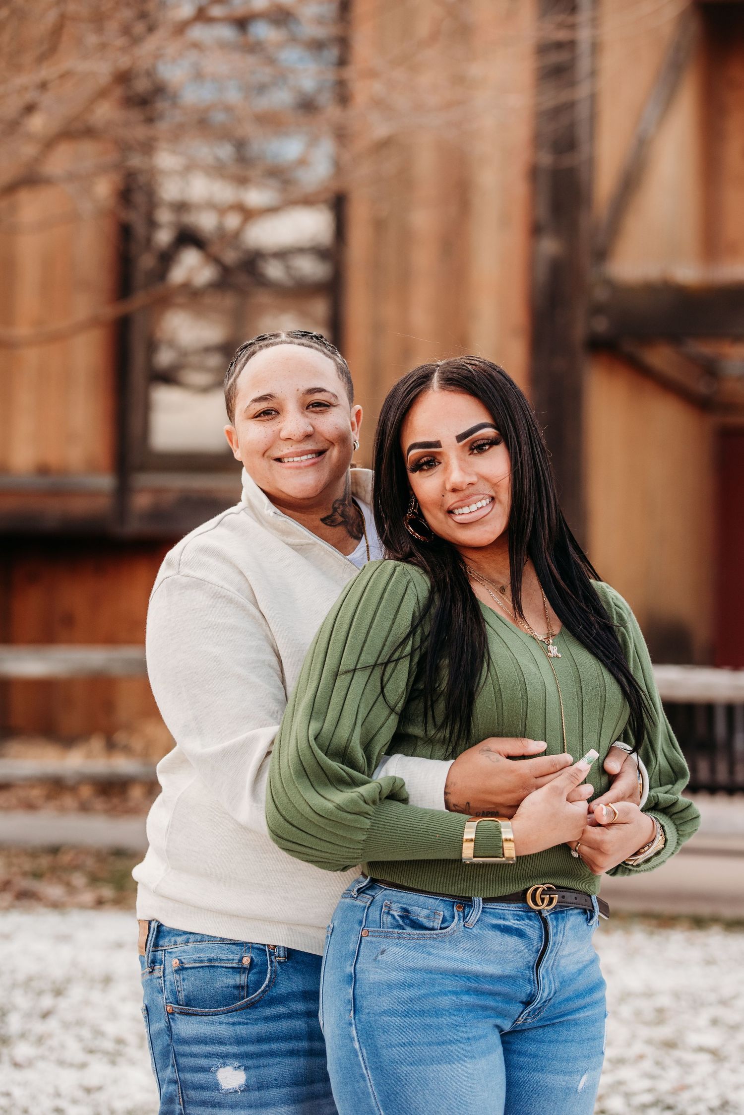 A couple in casual attire embracing while standing outdoors against a wooden building backdrop.