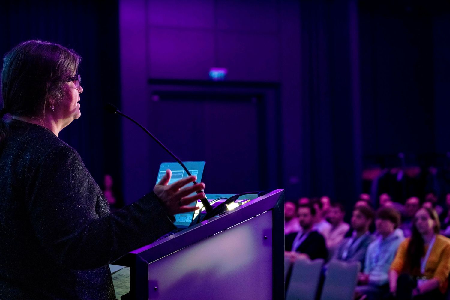 Speaker at a podium addresses a darkened conference hall with purple lighting.