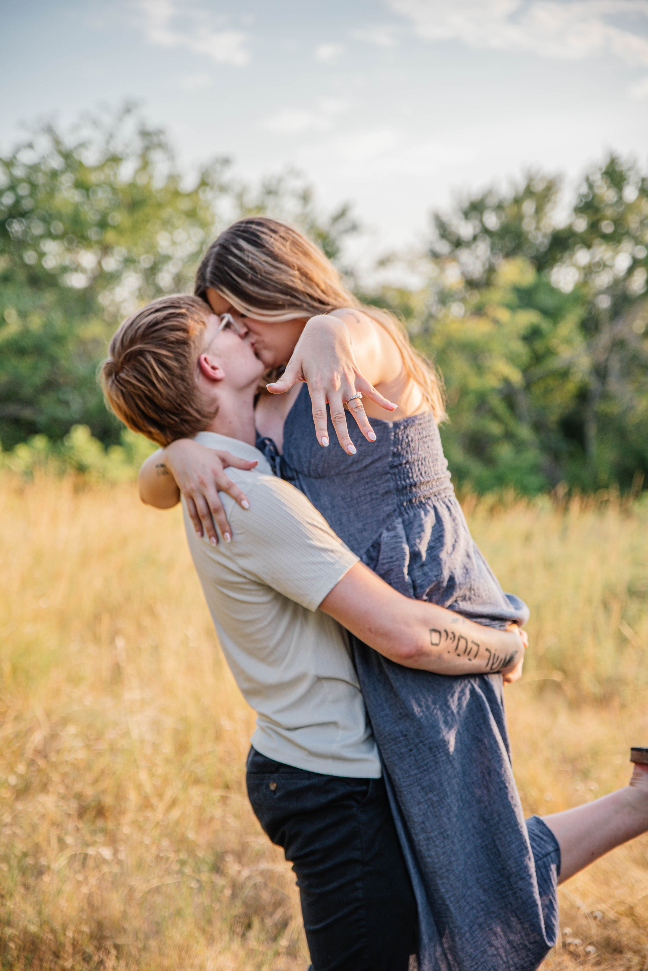 Couple sharing a romantic kiss in a golden field, man lifting woman in gray dress outdoors.